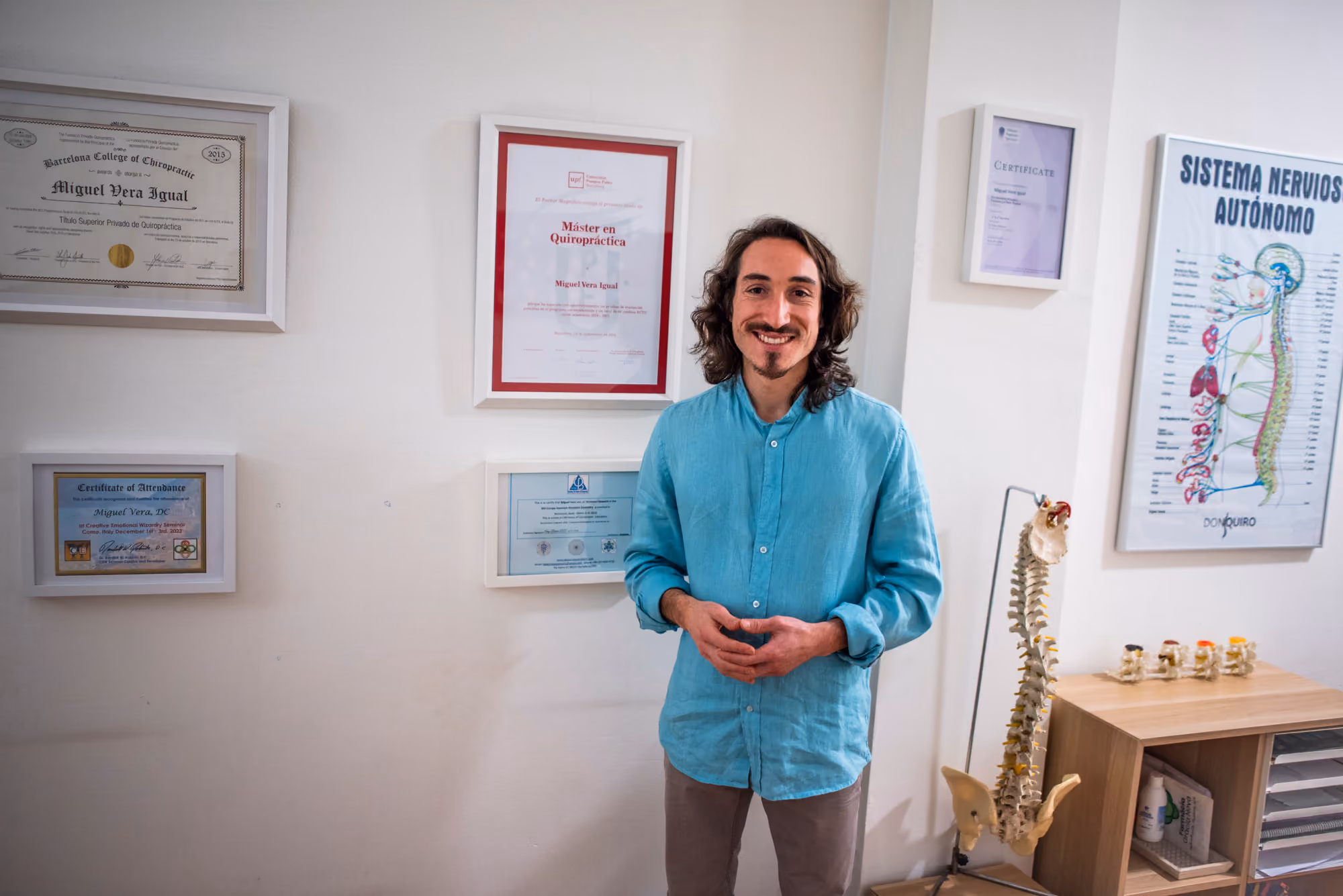 Miguel Vera, de pie y sonriente, delante de la pared con sus títulos y certificados enmarcados.
